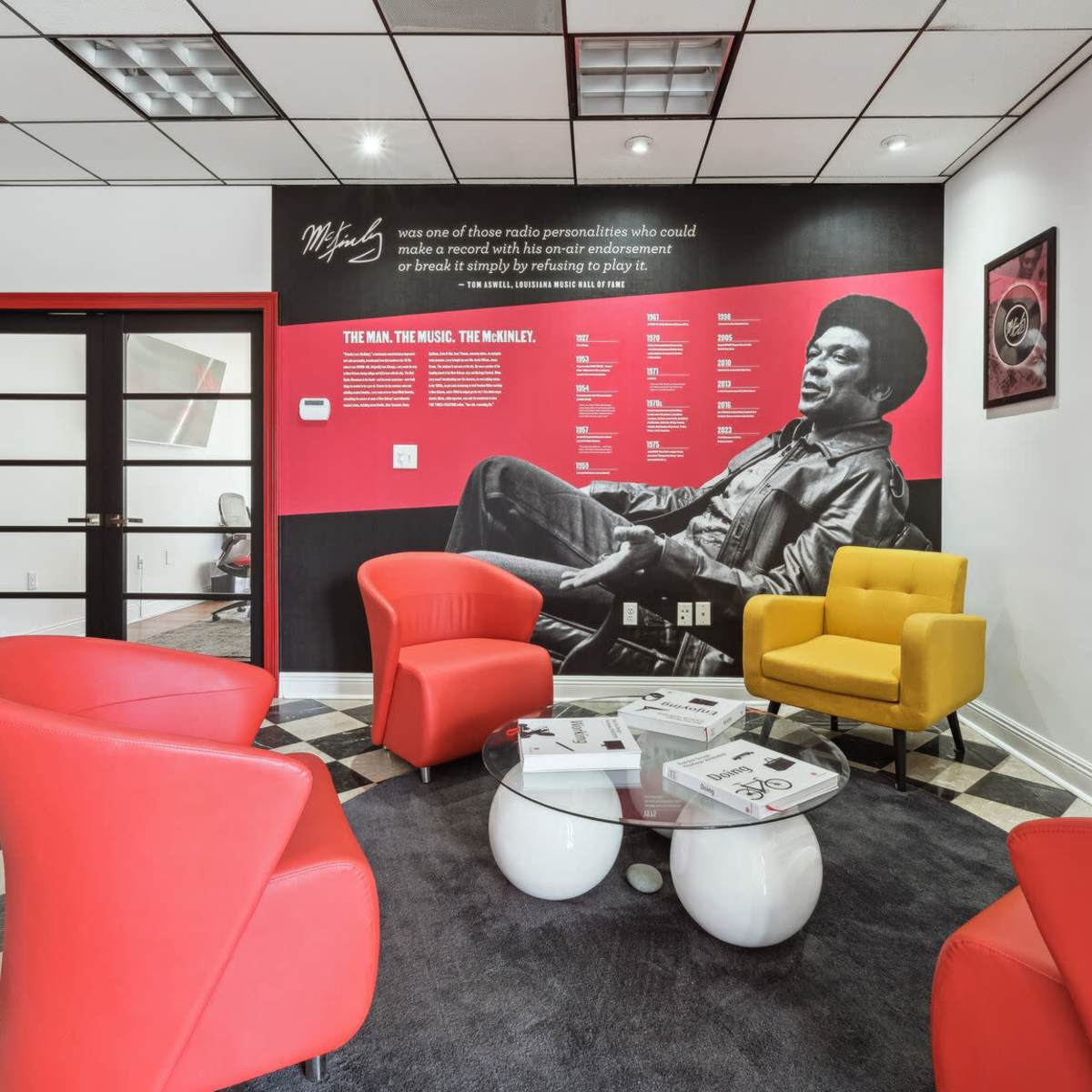 A modern lounge area with four colorful chairs and a table, alongside a wall featuring a large black-and-white mural of a musician and informational text.