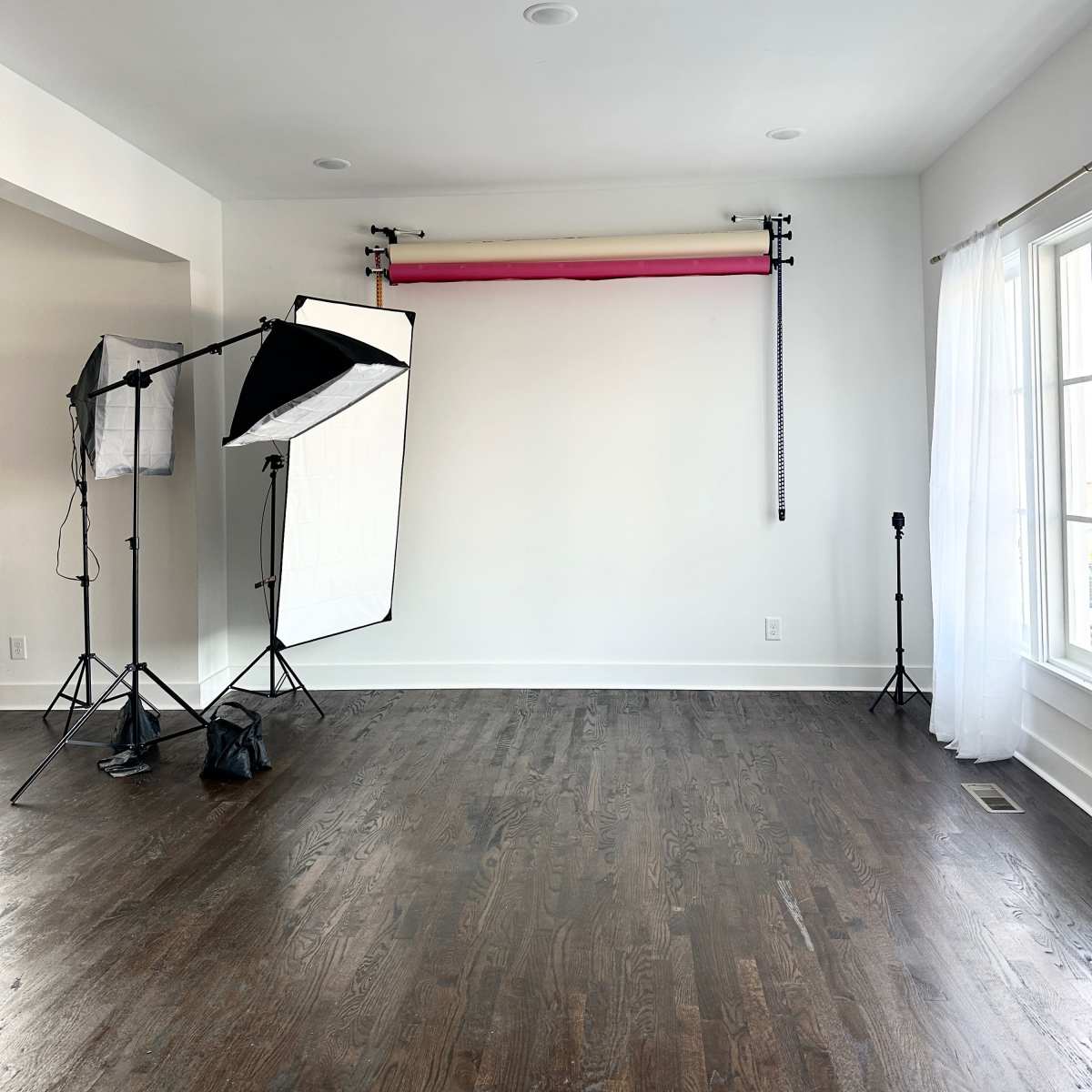 A vacant room with wooden flooring, two softbox light stands, a backdrop support system with a red and gray backdrop, and a single black stand.