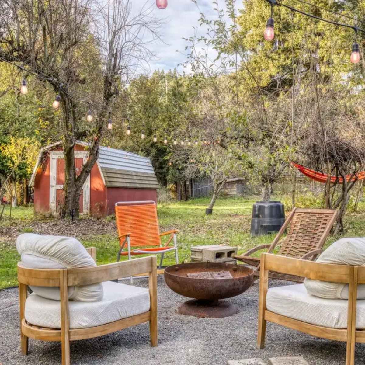 A backyard area with four outdoor chairs arranged around a circular fire pit, string lights overhead, and a red shed in the background.