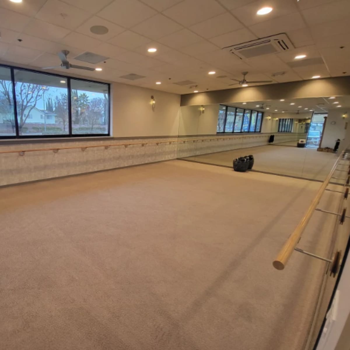 A spacious workout studio with large windows, a mirrored wall, and a wooden ballet barre along one side.