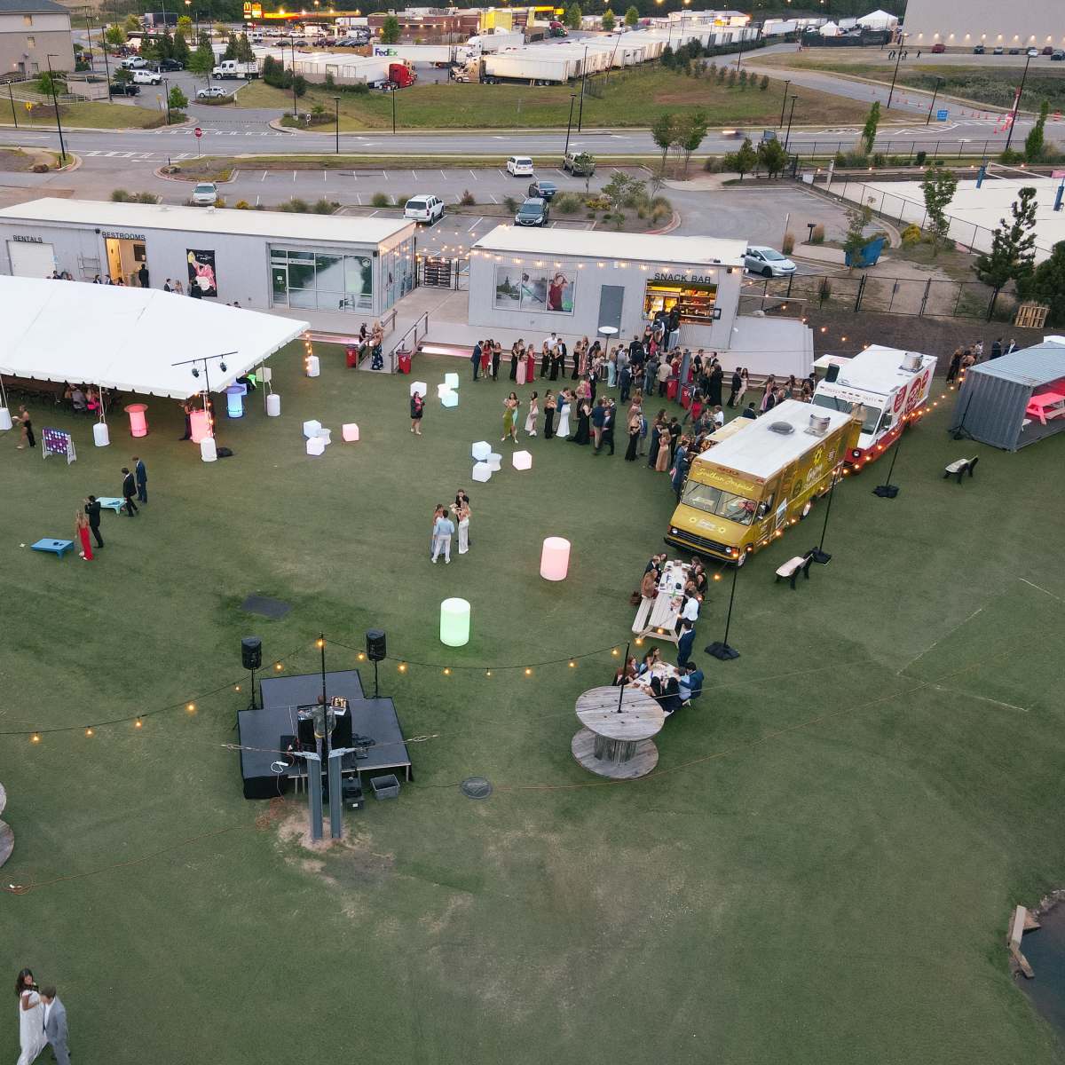 An outdoor event is taking place on a large grassy area with a stage, food truck, and guests mingling near illuminated decorations.