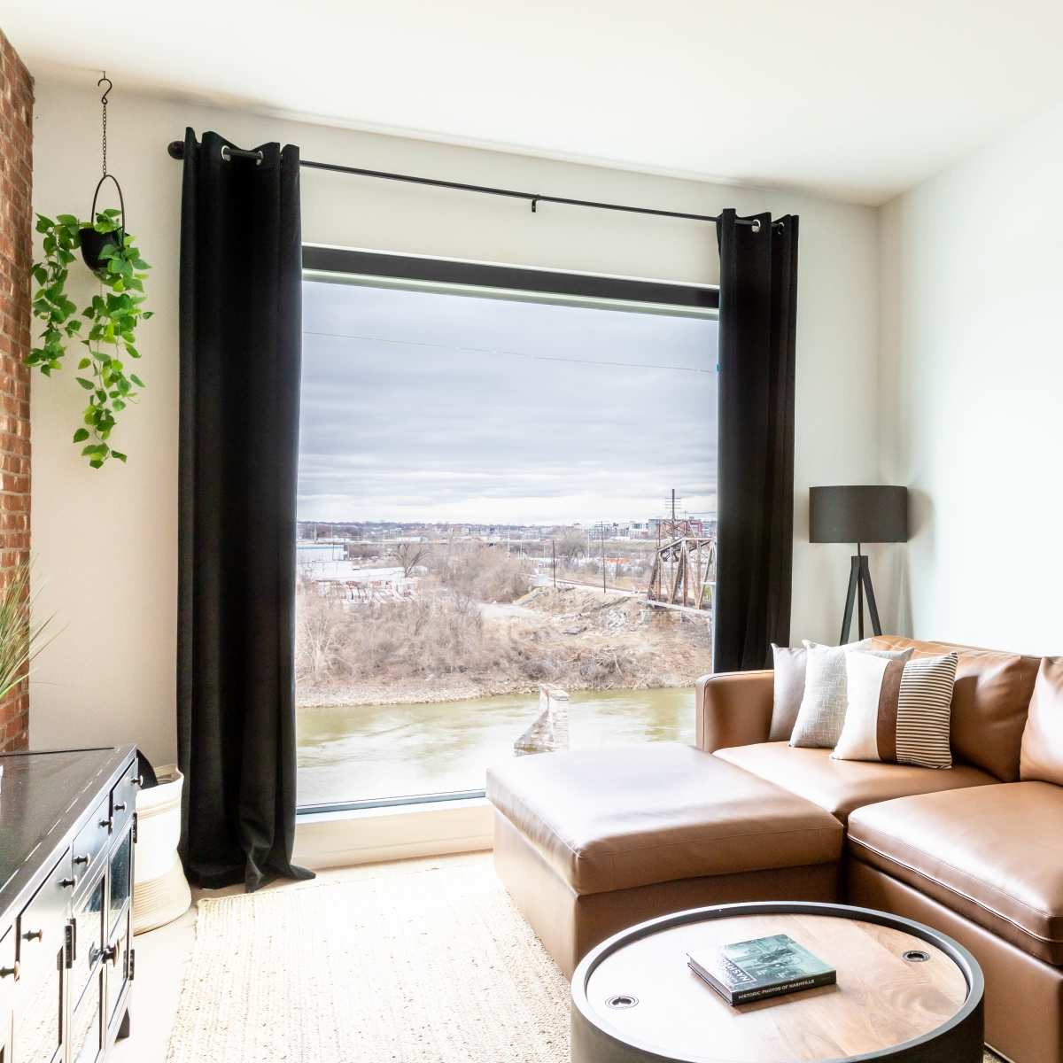 A modern living room features a large window overlooking a river, with a brown sectional sofa and a round coffee table.
