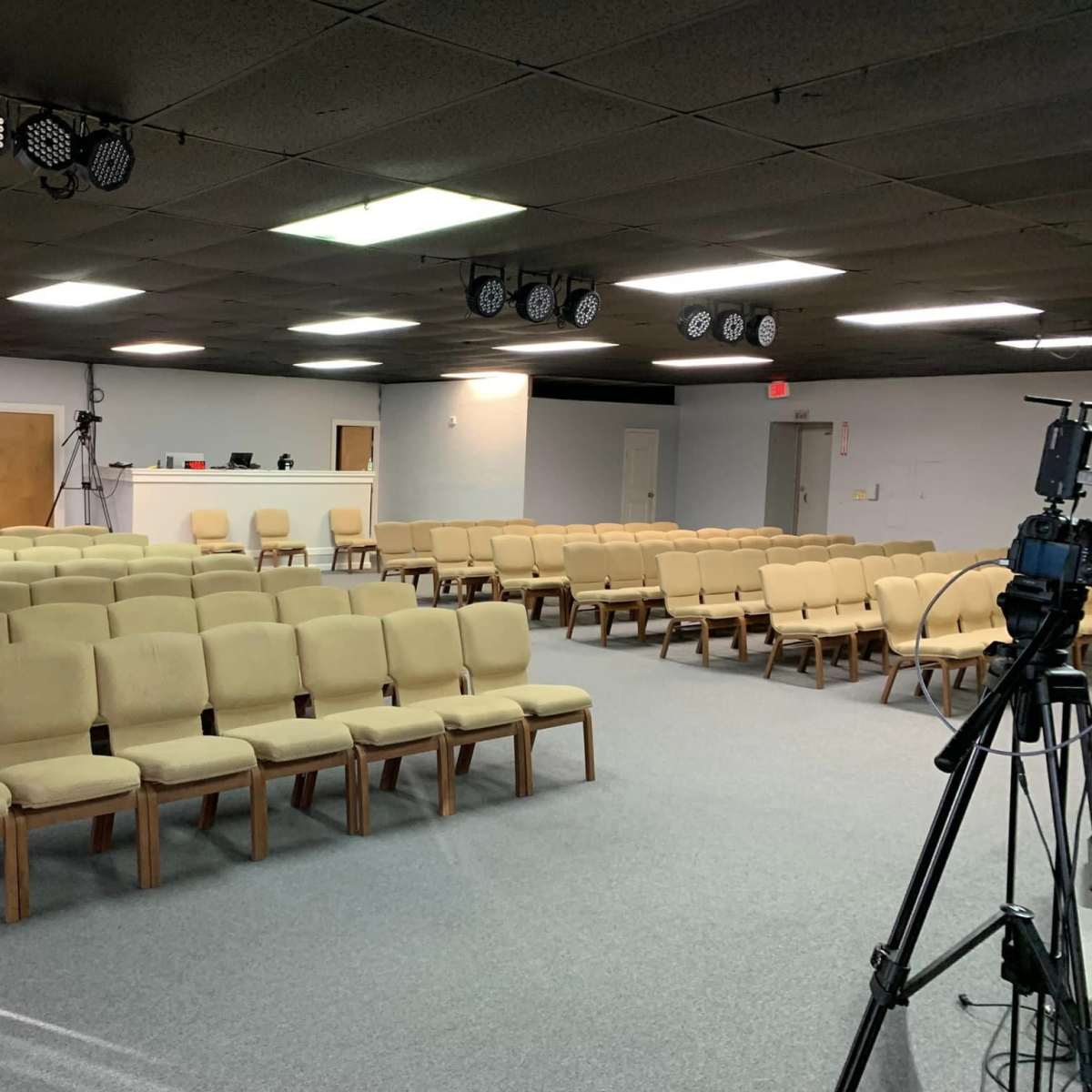A sparsely arranged seating area with beige chairs facing a blank wall in a room with a low ceiling, equipped with spotlights and cameras.