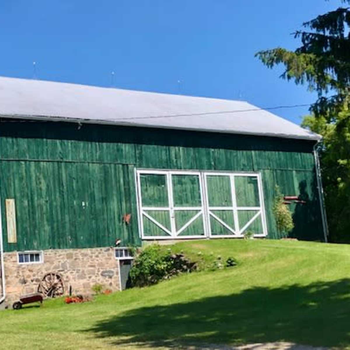 A large green barn with a stone foundation sits on a grassy slope surrounded by trees.