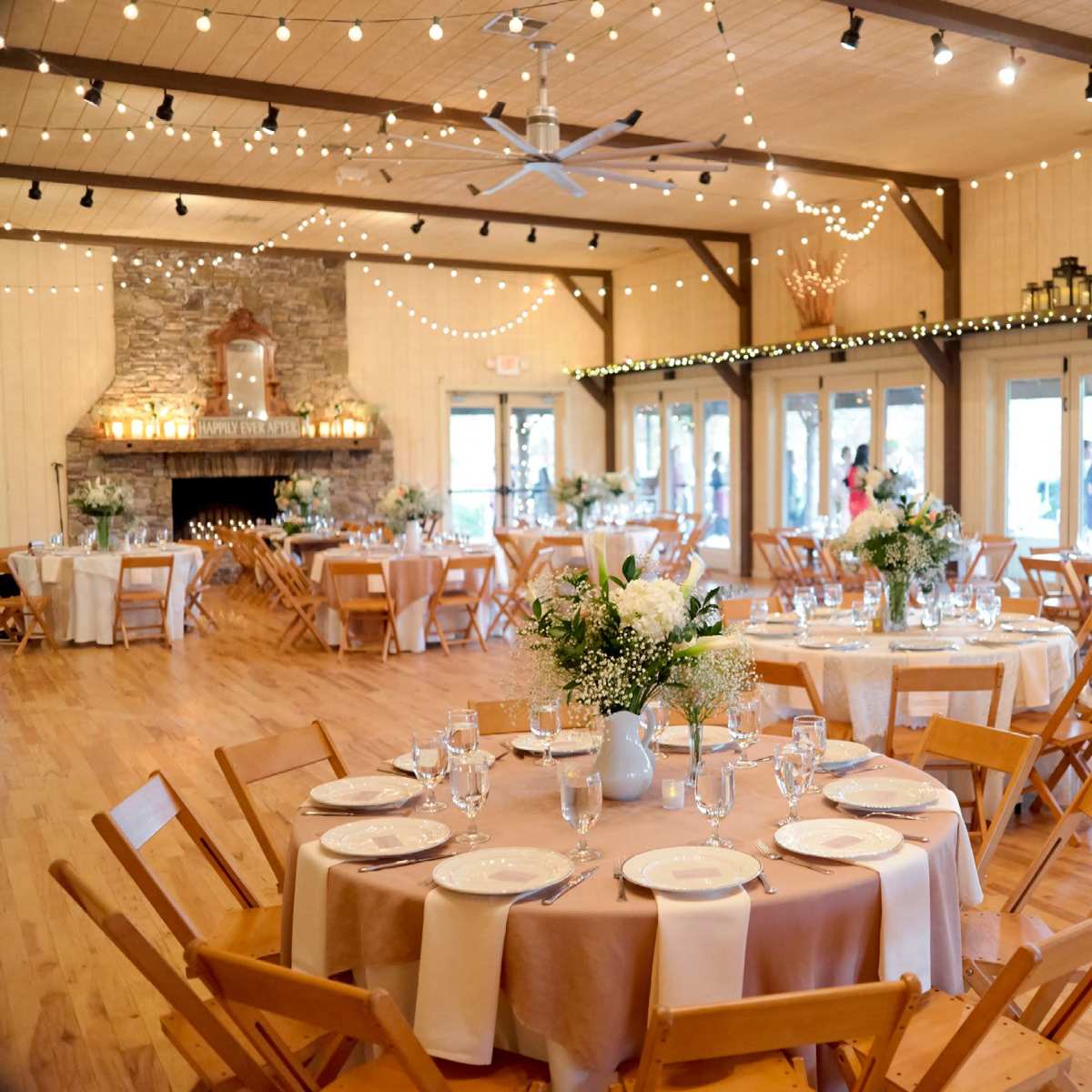 A well-decorated banquet hall with round tables set for a formal event, featuring floral centerpieces and string lights overhead.