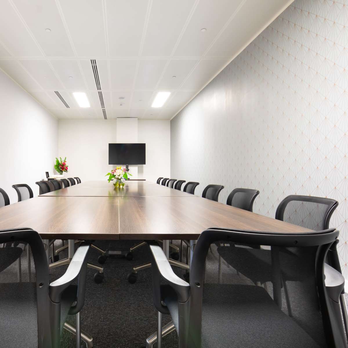 A large conference room features a rectangular wooden table surrounded by black mesh chairs, with a television mounted on the wall and decorative elements in the background.