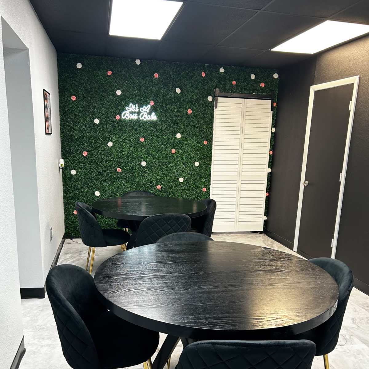 A modern room with dark round tables and black chairs, set against a green wall adorned with white flowers and a neon sign.
