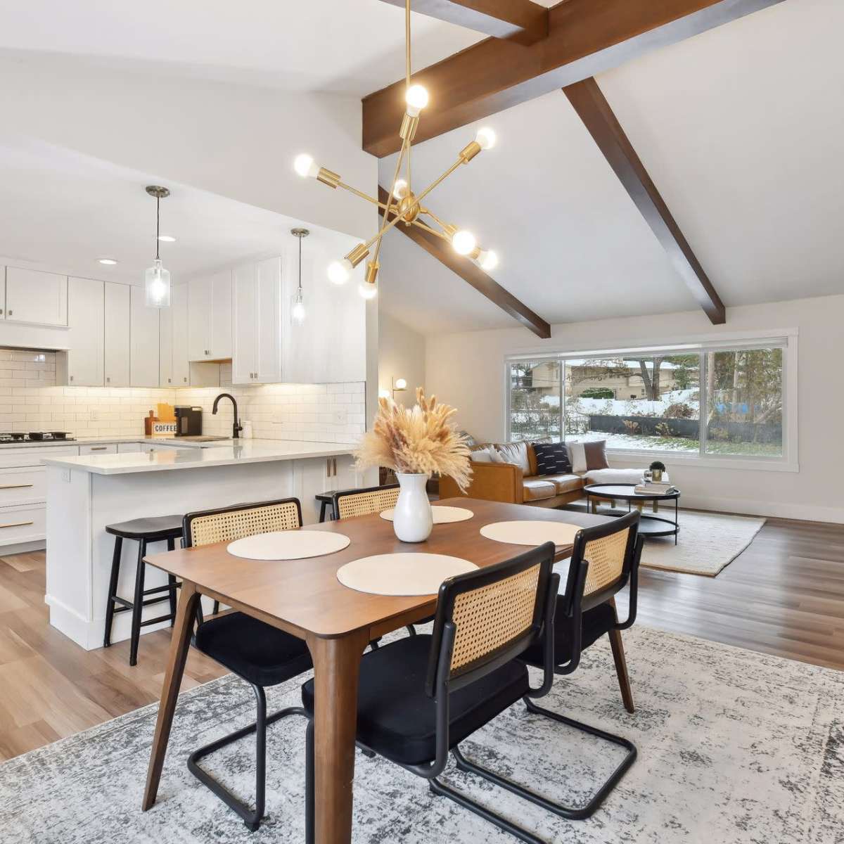 A modern living space features a dining area with a table and chairs, a kitchen with white cabinetry, and a cozy sitting area illuminated by natural light through large windows.
