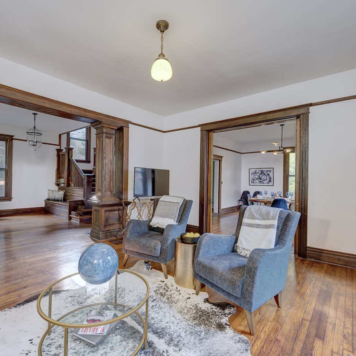 A spacious living area features hardwood floors, two blue armchairs, a glass coffee table, and a staircase leading to the upper level.