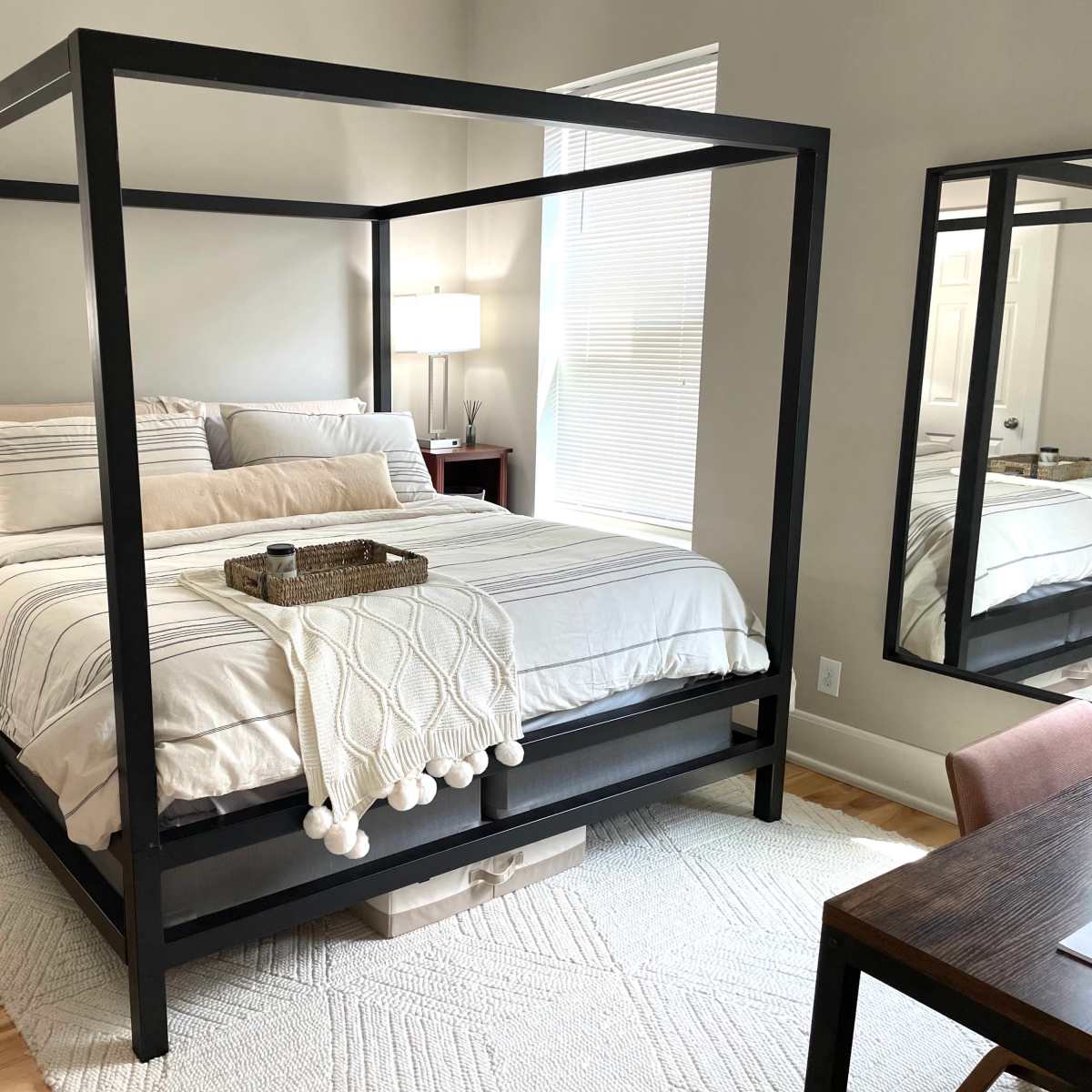 A modern bedroom featuring a four-poster bed with a light-colored comforter, two bedside lamps, and a mirror reflecting a desk area.