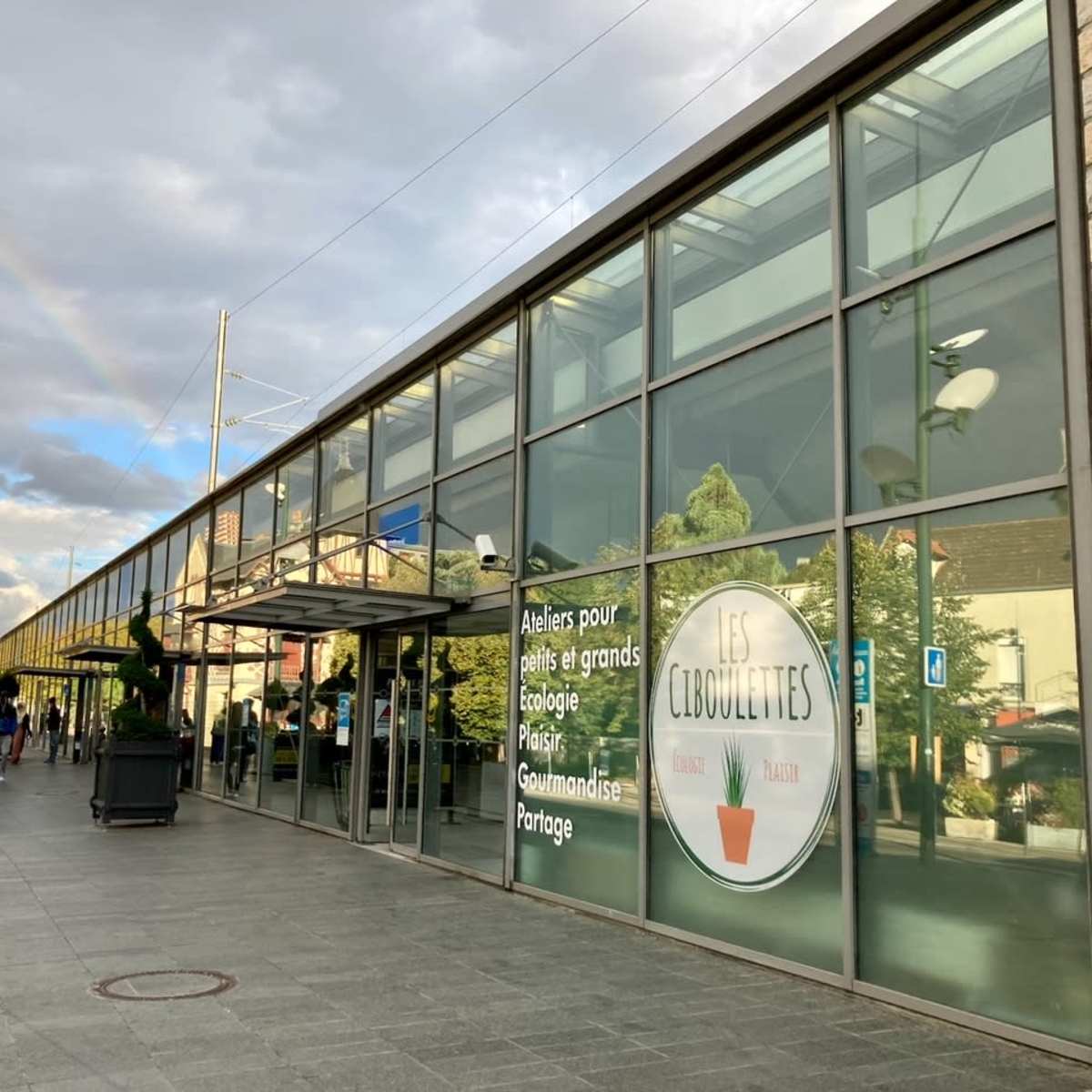 A large, glass-fronted building with a sign reading "Les Libouclettes," surrounded by planters and a rainbow in the background.