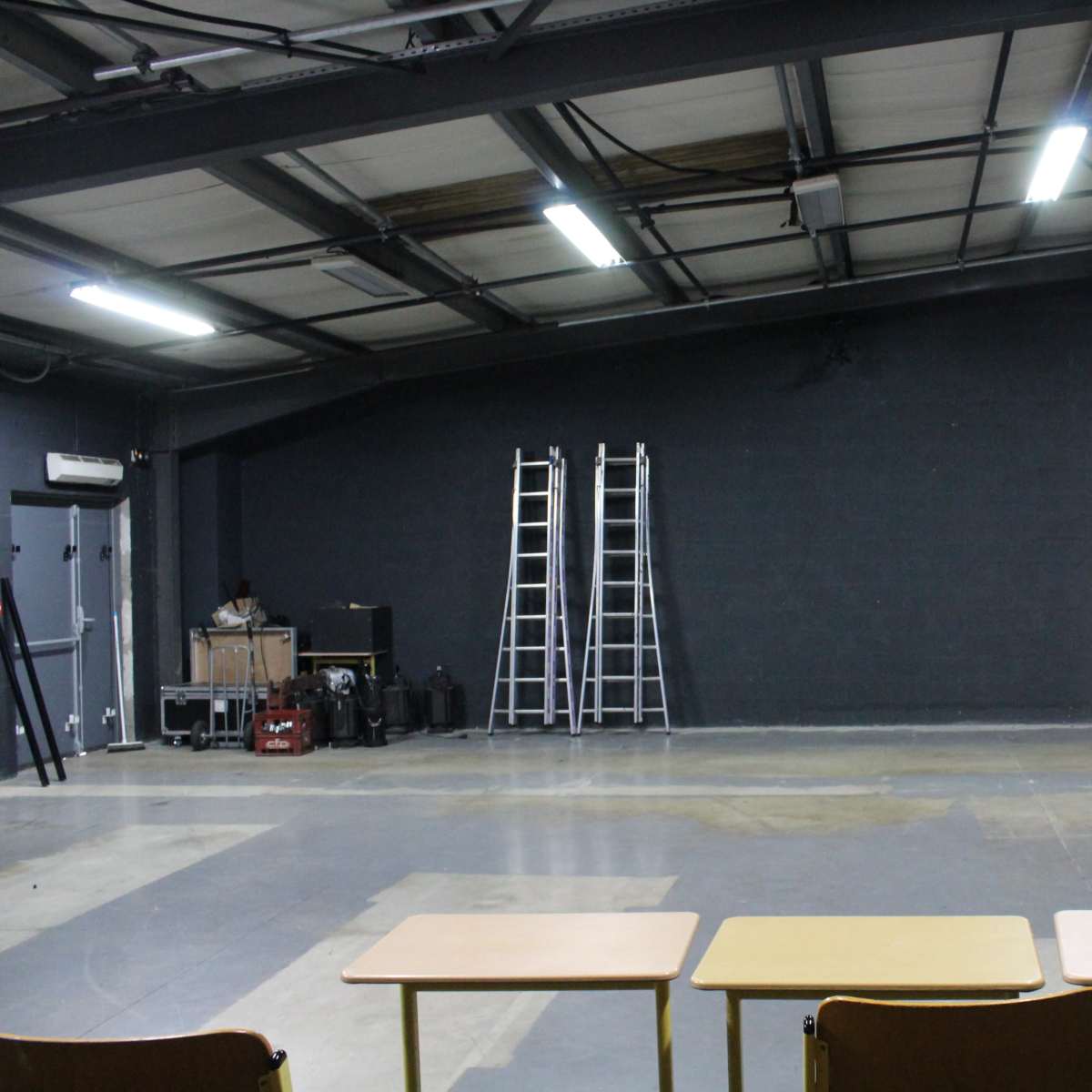 An empty industrial space with three tables in the foreground, two ladders leaning against a wall, and various equipment in the background.