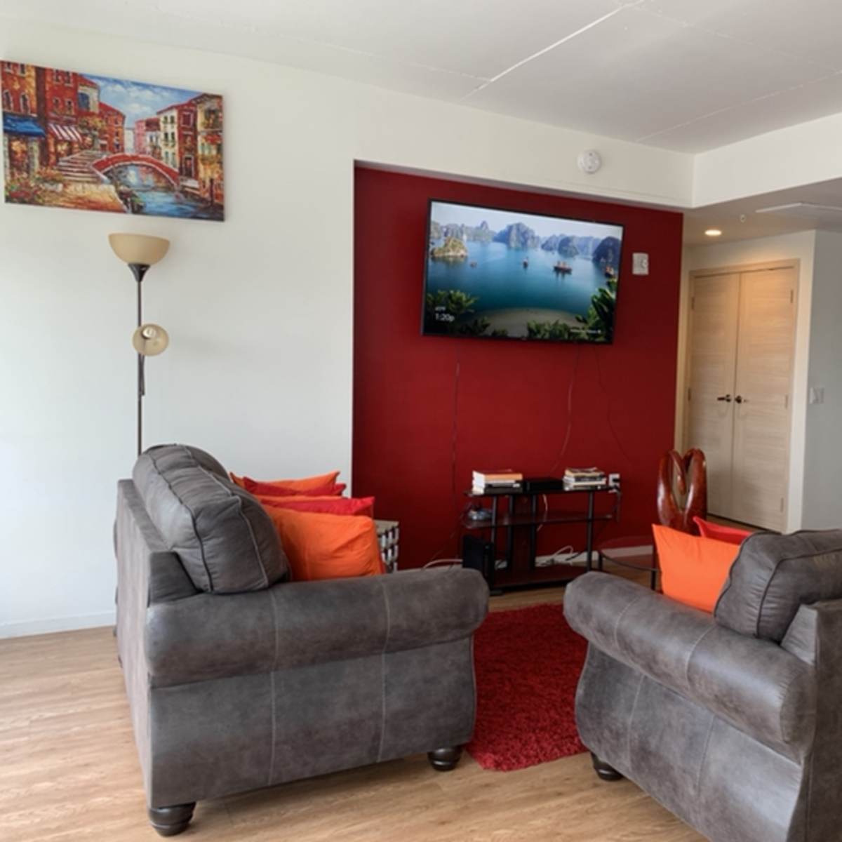 A living room with two gray sofas facing a wall-mounted television, a red accent wall, and a piece of artwork depicting a scenic view.
