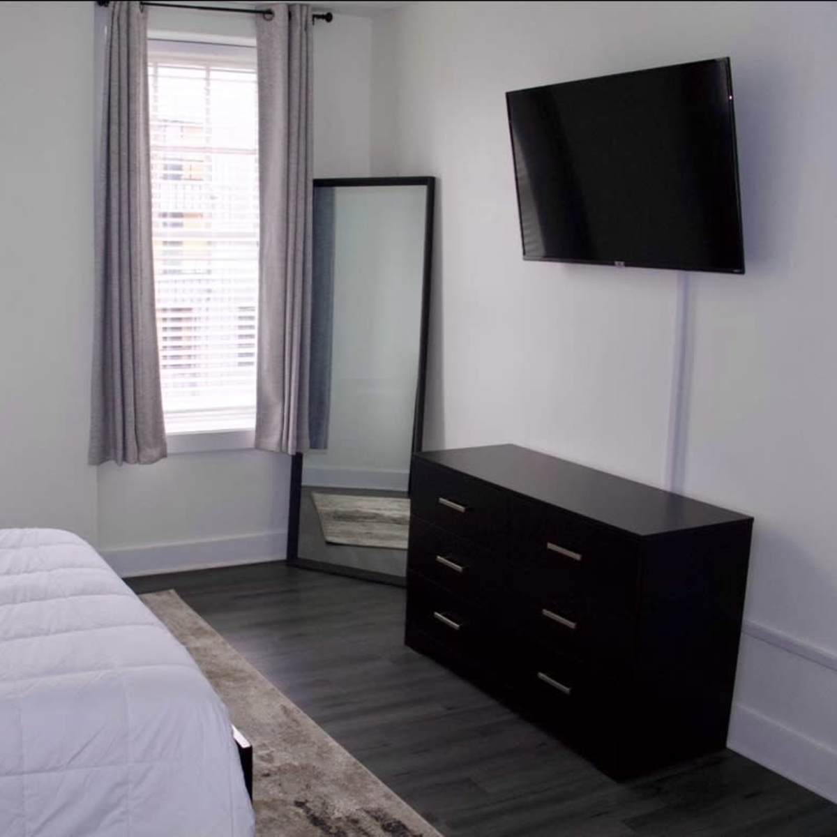 A bedroom features a bed covered with a white comforter, a dresser with five drawers, a full-length mirror, and a mounted television on the wall, all set against white walls and gray curtains.