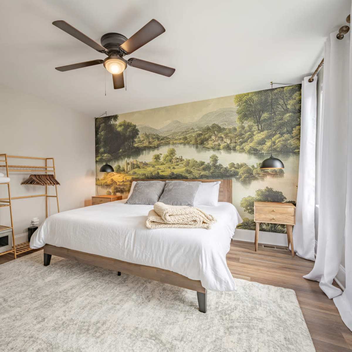 A bedroom features a queen-sized bed with a scenic mural of a landscape, complemented by a ceiling fan and minimalist decor.