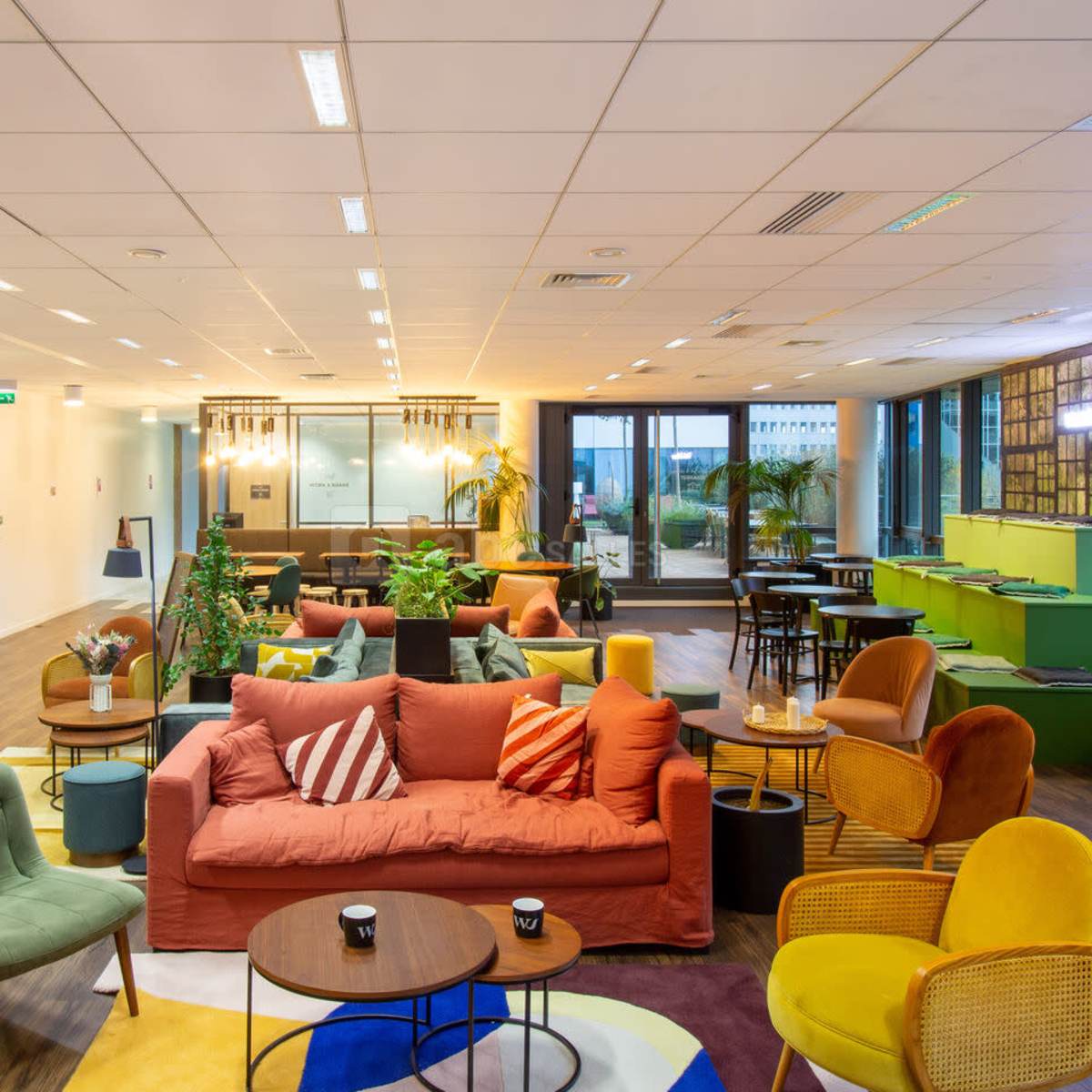 A modern office lounge area featuring a variety of seating arrangements, plants, and large windows that allow natural light to enter.