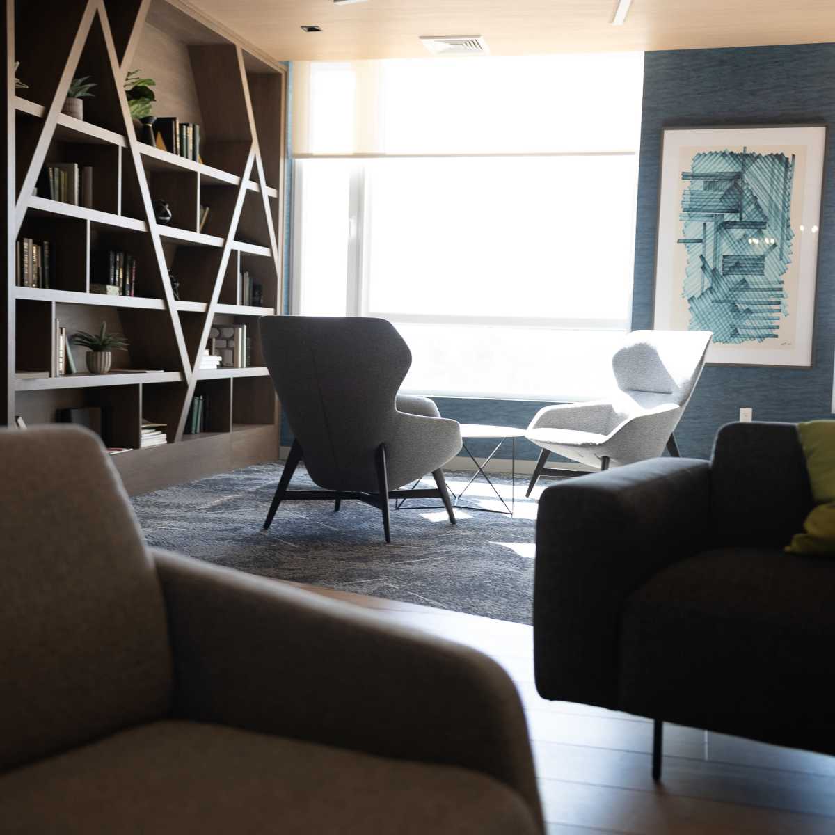 A modern lounge area with two armchairs and a bookshelf against a wall, illuminated by natural light from large windows.