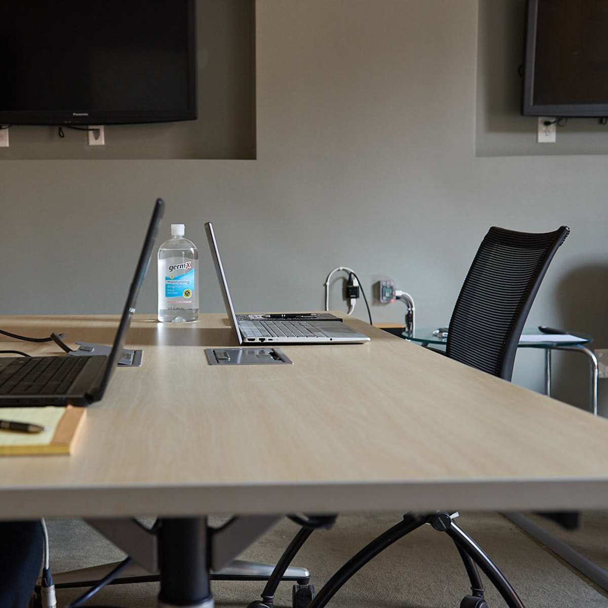 A conference room table is set up with two laptops, a notepad, a pen, and a bottle of water, with two black chairs positioned at the table.