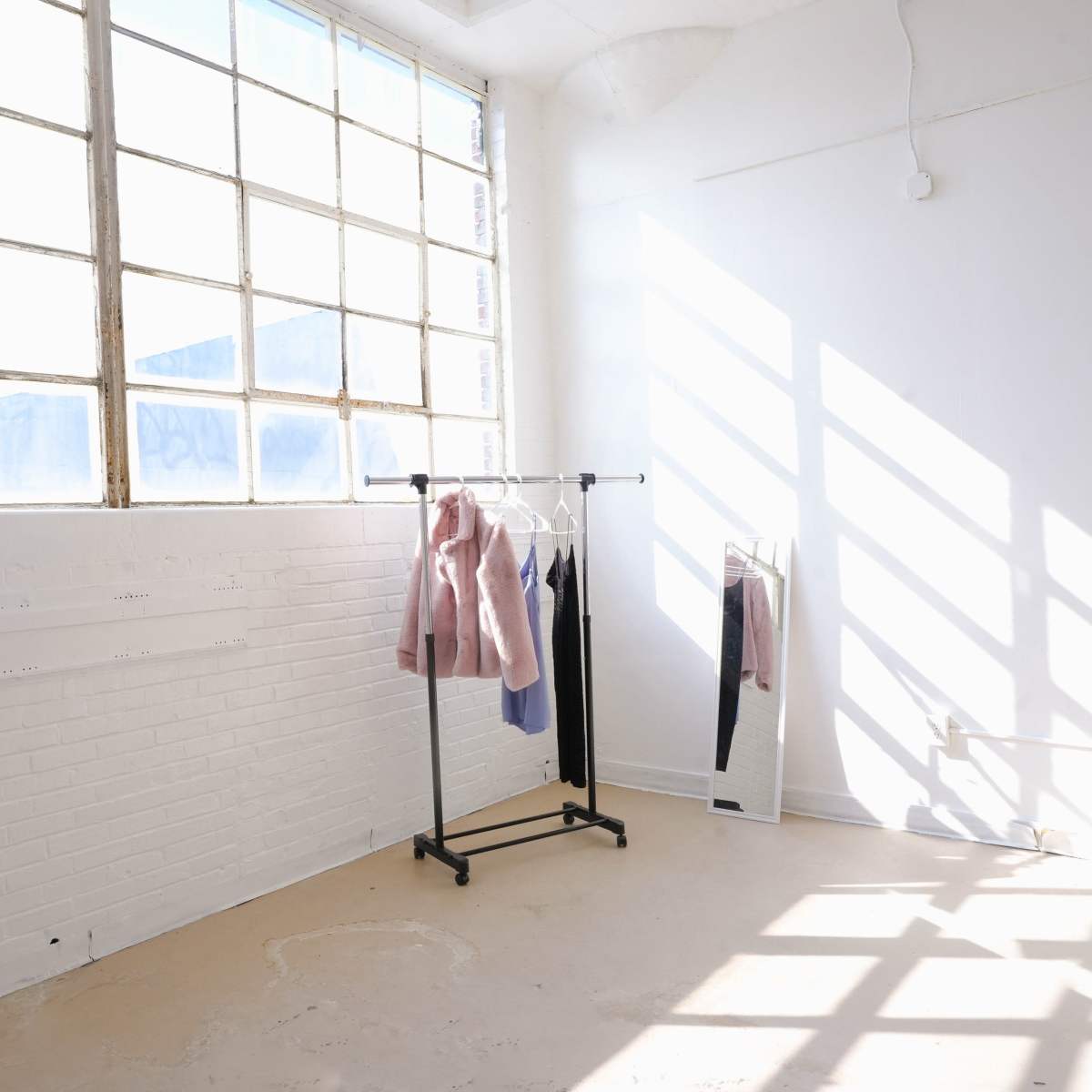A bright, minimalistic room features a clothing rack with a few garments and a full-length mirror against a wall with large windows.