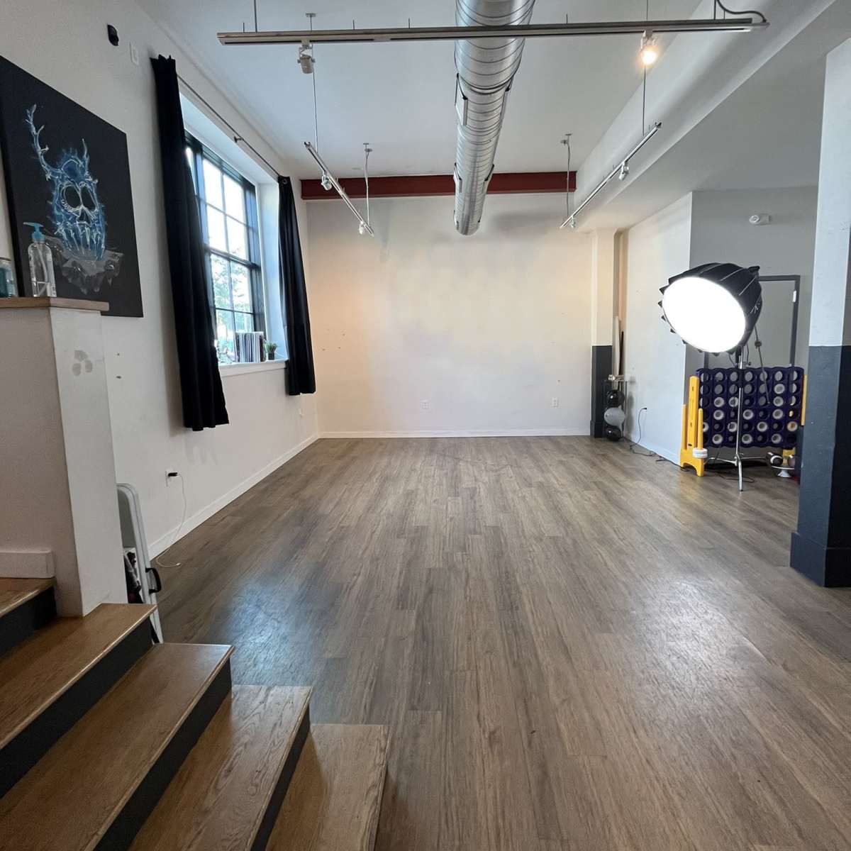 A spacious, empty studio with wooden flooring, large windows, and a spotlight set up in the corner.