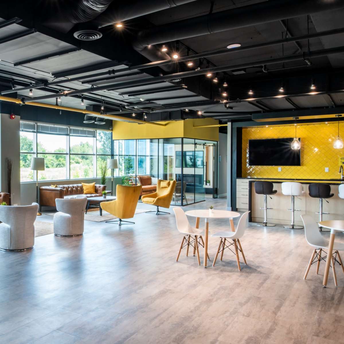 A modern office lounge area with various seating options, large windows, and a kitchen space in the background.