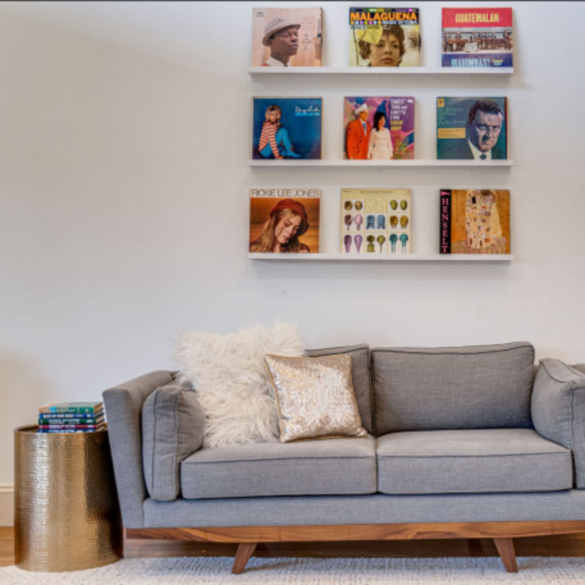 A gray sofa with decorative pillows is positioned in front of a wall displaying shelf-mounted vinyl records.