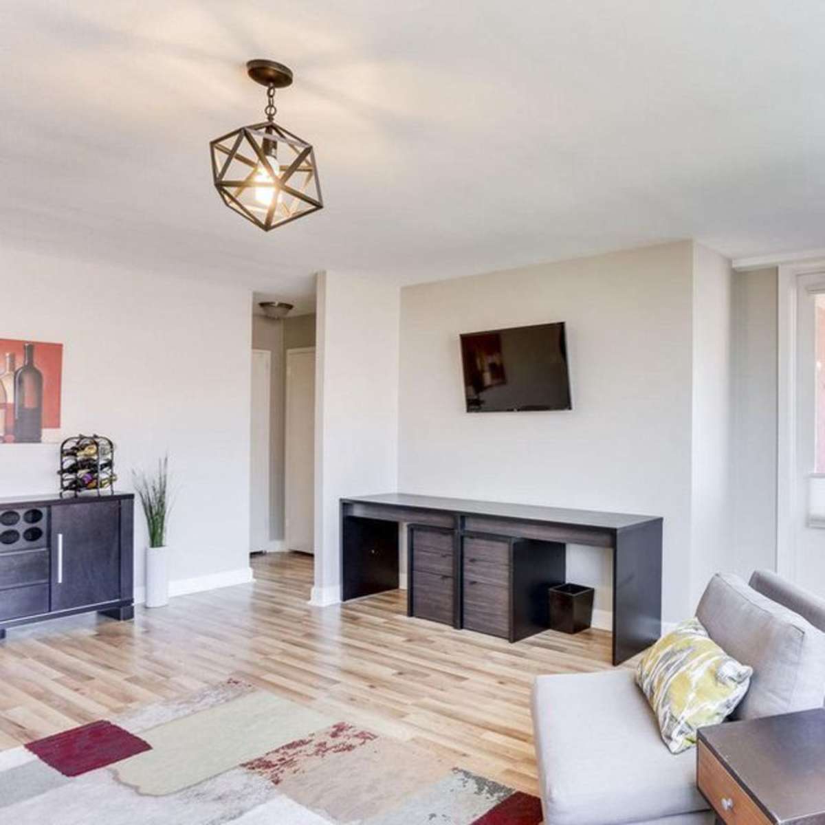 A modern living room featuring hardwood floors, a wall-mounted TV, a light fixture hanging from the ceiling, and a sofa with a decorative pillow.