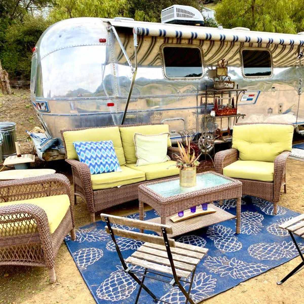 An Airstream trailer is parked next to a seating area with wicker chairs and a table surrounded by decorative items.