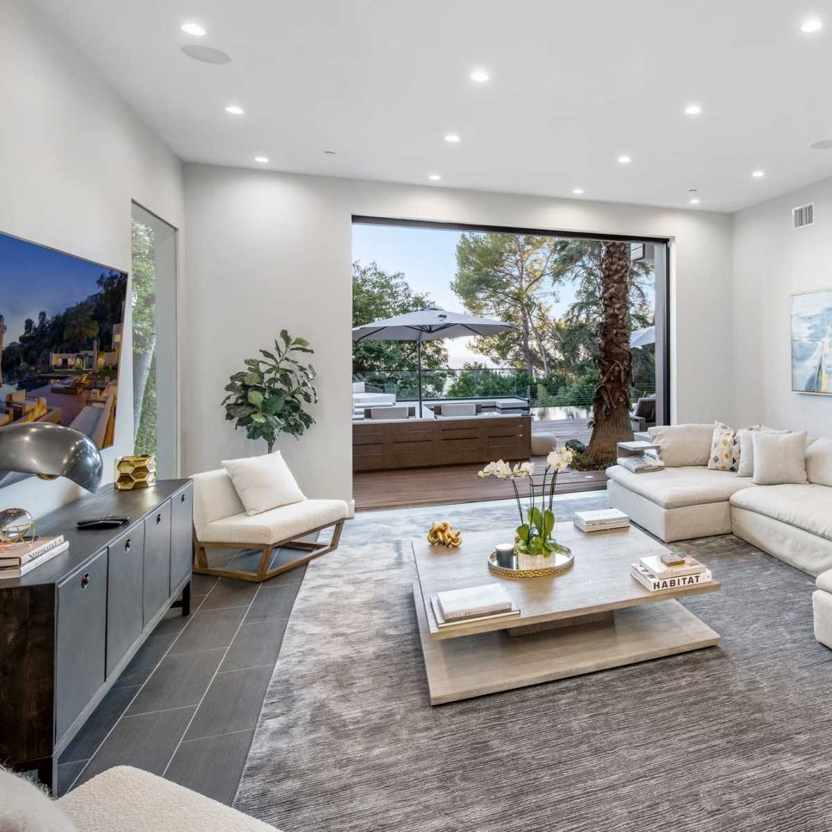 A modern living room features a large sectional sofa, a coffee table, and floor-to-ceiling windows opening to an outdoor patio with a pool.
