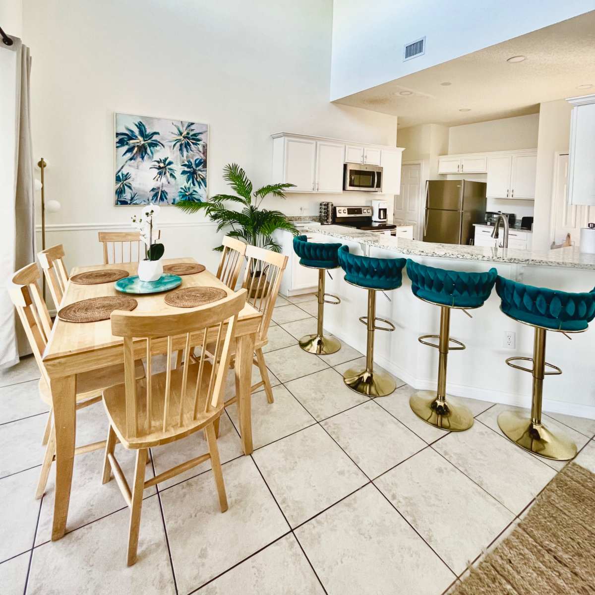 A modern kitchen and dining area feature a wooden table with four chairs, turquoise bar stools at a counter, and a decorative plant.