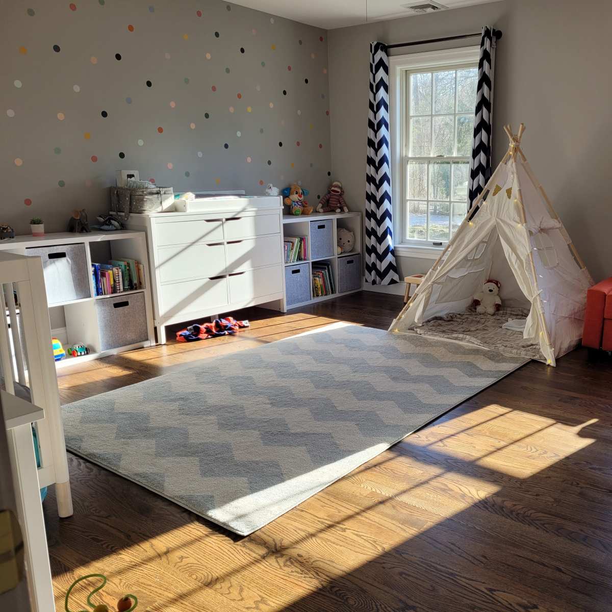 The room features a playful design with a sofa, a teepee tent, and organized storage, illuminated by natural light through large windows.