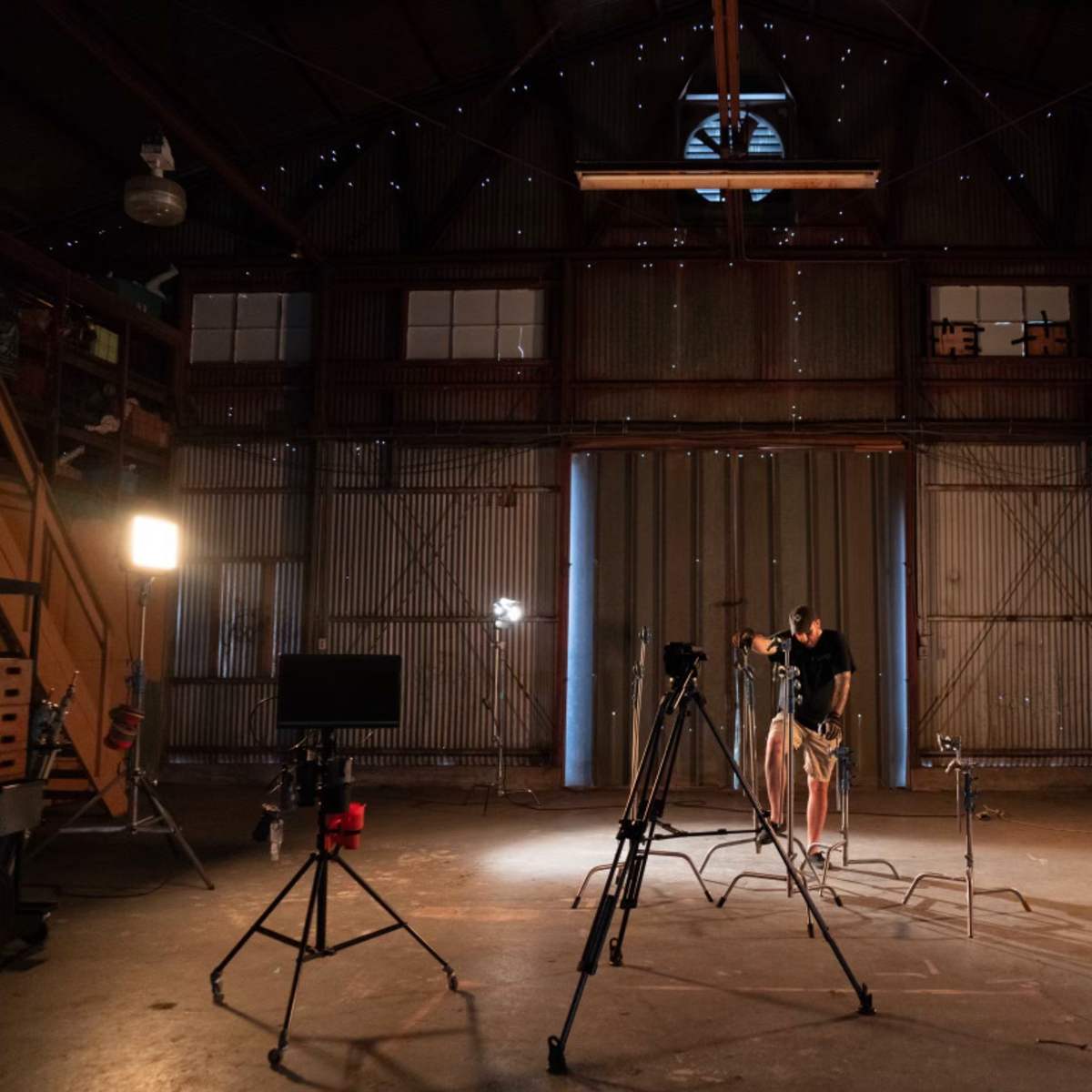 A studio setup is visible in a barn-like space, featuring camera equipment, lighting fixtures, and a person preparing for a shoot.