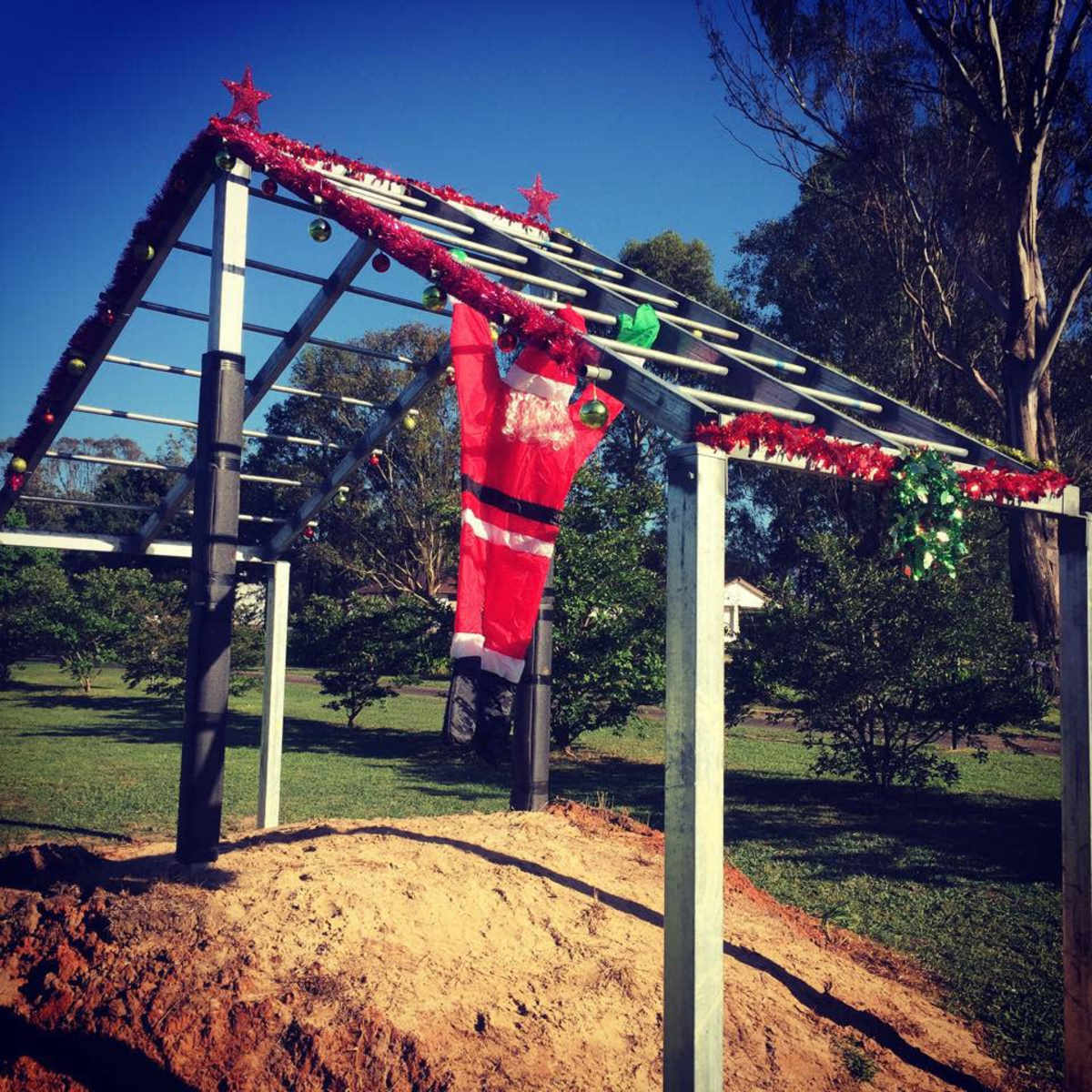 A metal frame decorated with holiday ornaments and a Santa Claus figure hangs in a grassy area surrounded by trees.