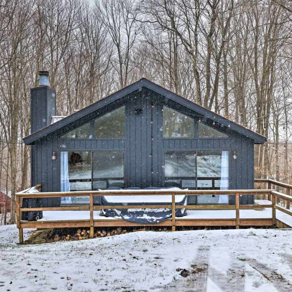 A dark blue cabin with large windows is situated in a snowy forest, featuring a wooden deck and a path leading up to it.