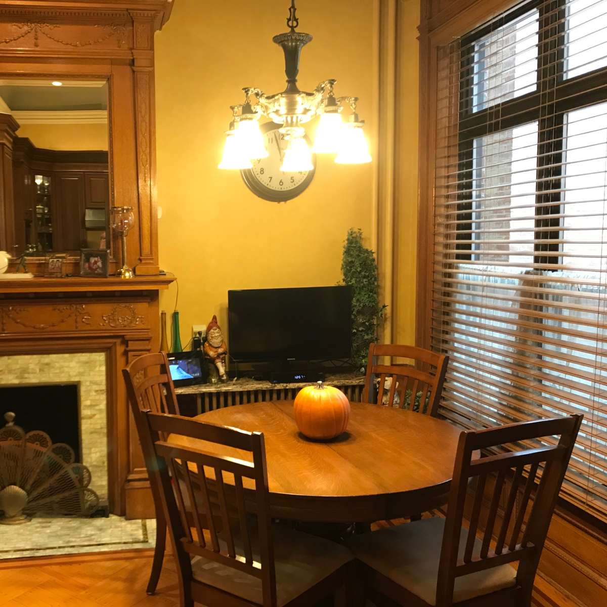 A wooden dining table with four chairs is positioned near a fireplace and a television in a warmly lit room.