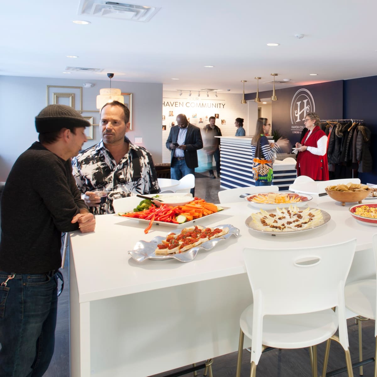 A social gathering in a modern space with people engaging in conversation around a white table filled with various food platters.
