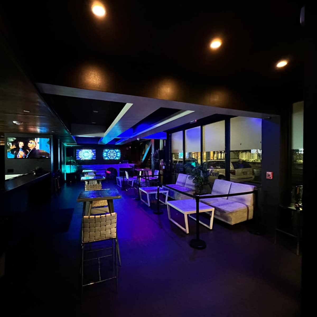 A modern lounge features comfortable seating, illuminated by colorful lights, with a bar area and screens displaying visuals.