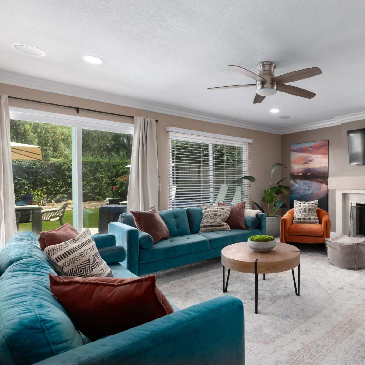 The living room features a turquoise sectional couch, two orange armchairs, a round coffee table, and a fireplace, with large windows overlooking a garden.