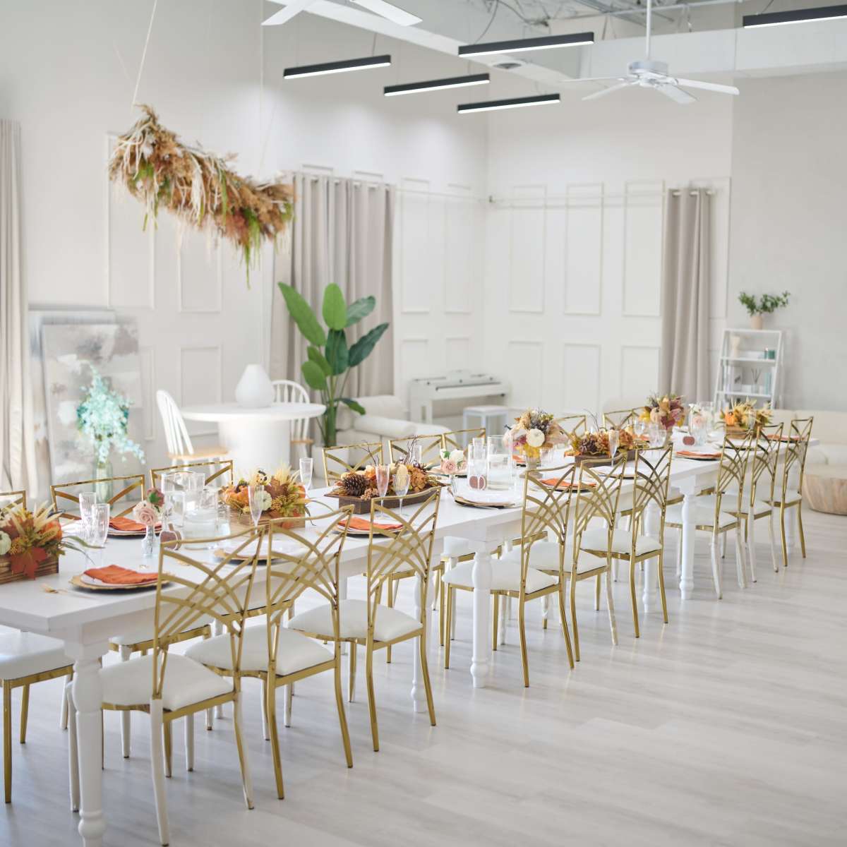 A long dining table is set with colorful centerpieces and tableware in a bright, modern event space with minimalistic decor and large windows.