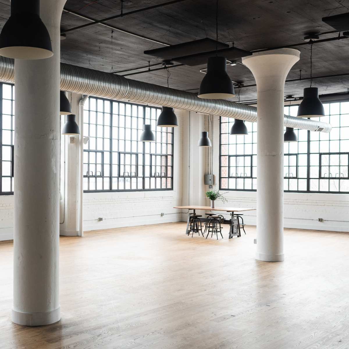 A spacious, industrial-style room with large windows, high ceilings, and a wooden floor, featuring several columns and a dining table with chairs.