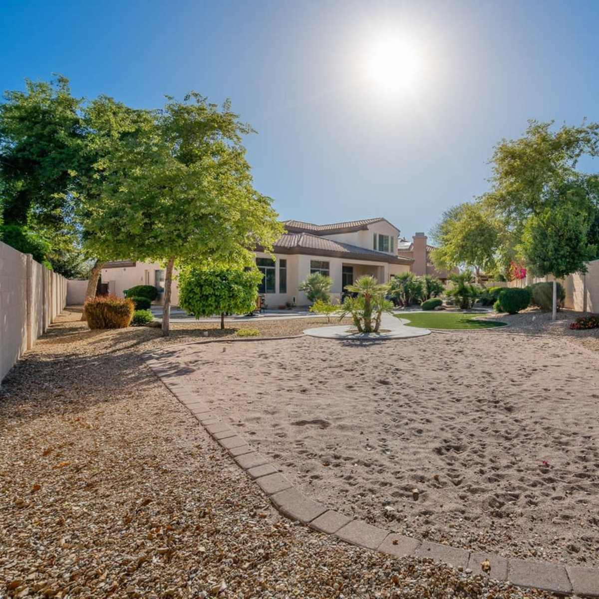 A spacious backyard with a sandy area surrounded by desert landscaping and a two-story house in the background under a bright sun.