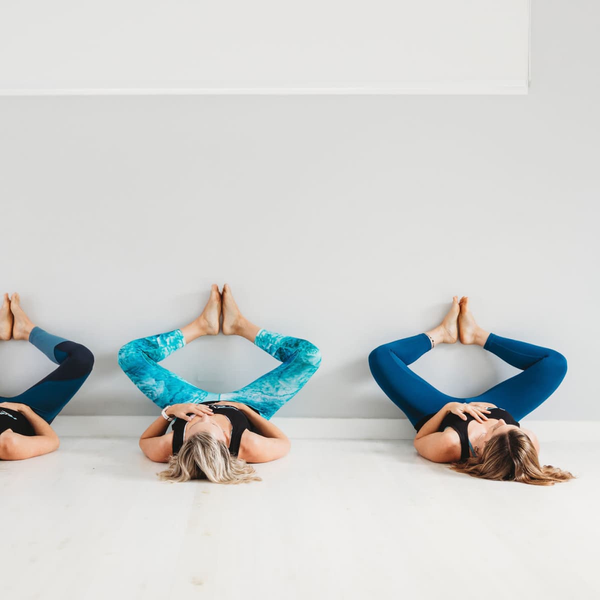Three individuals perform a yoga pose on a hardwood floor, with their legs raised against a wall.