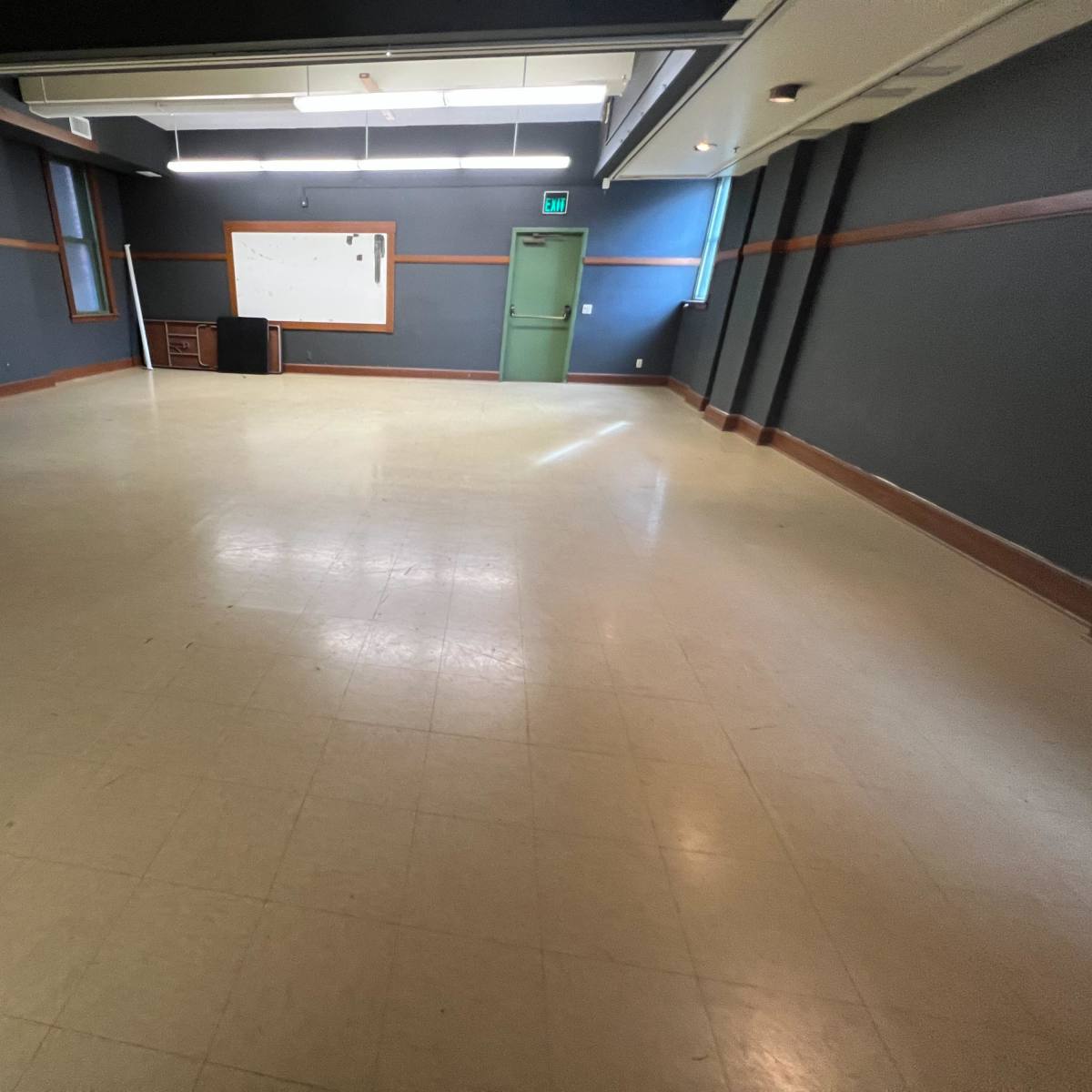 An empty classroom with a whiteboard, a desk in the corner, and green doors under overhead lights.