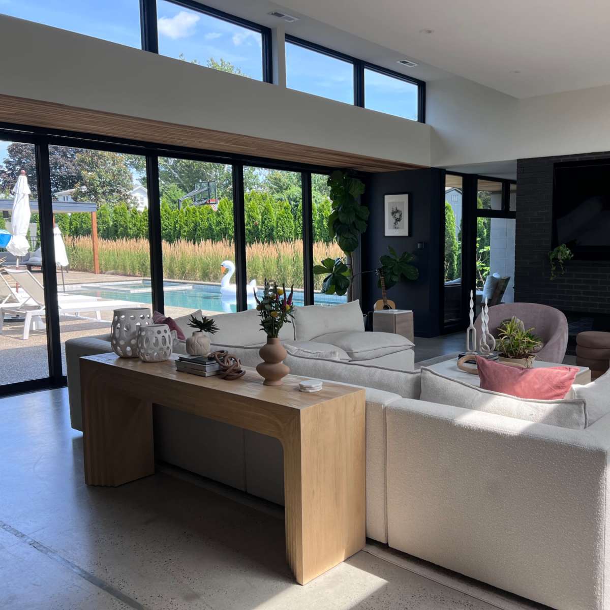 A modern living room features a light-colored sofa, a wooden coffee table, and large glass doors that open to a pool area surrounded by greenery.