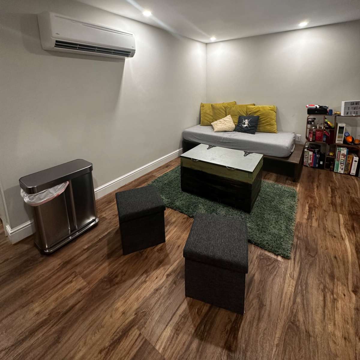 A cozy living space featuring a gray sofa with yellow throw pillows, a wooden coffee table, two black stools, a trash can, and a green rug on a hardwood floor, all under recessed lighting.