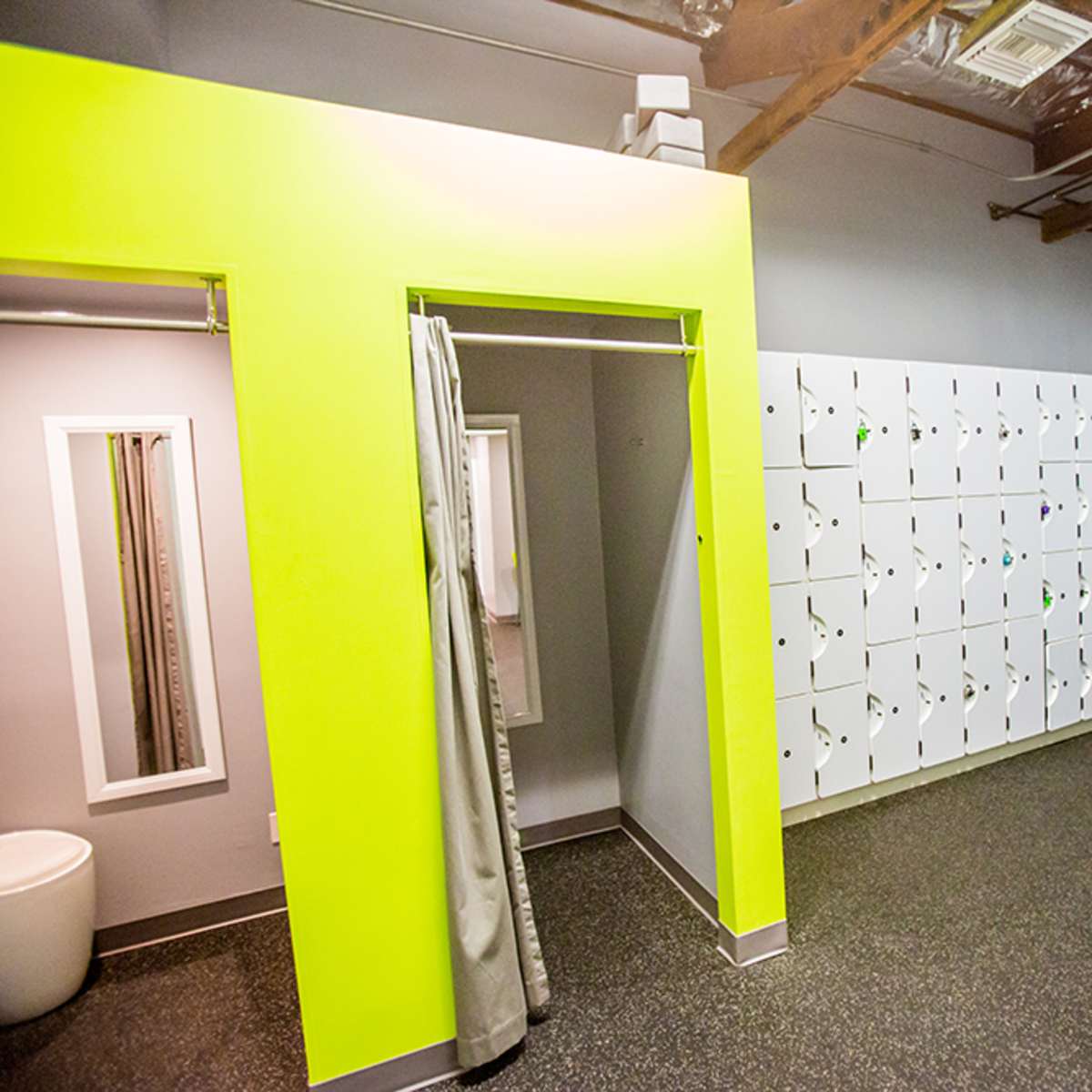 A modern changing room featuring a bright green accent wall, a row of white lockers, a shower stall, and a chair.