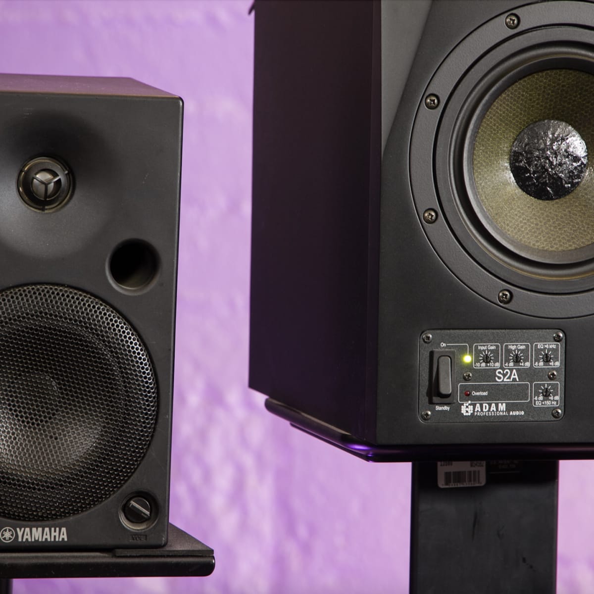 Two studio monitor speakers, one branded Yamaha on the left and another branded Adam on the right, positioned against a purple wall.