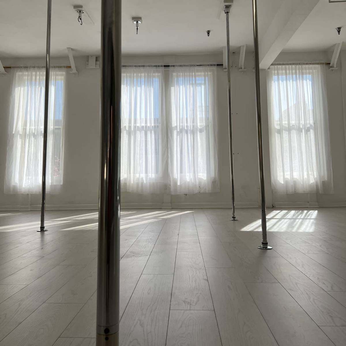 A spacious room with polished wooden floors and several vertical poles, illuminated by natural light streaming through sheer curtains.