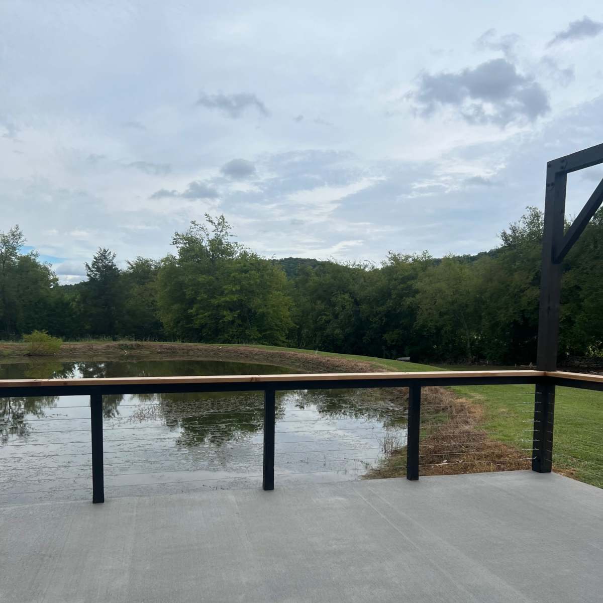 A concrete patio overlooks a tranquil pond surrounded by greenery and trees.