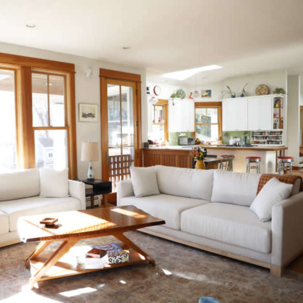 A bright living room features two white sofas arranged around a wooden coffee table on a patterned rug, with large windows illuminating the space.